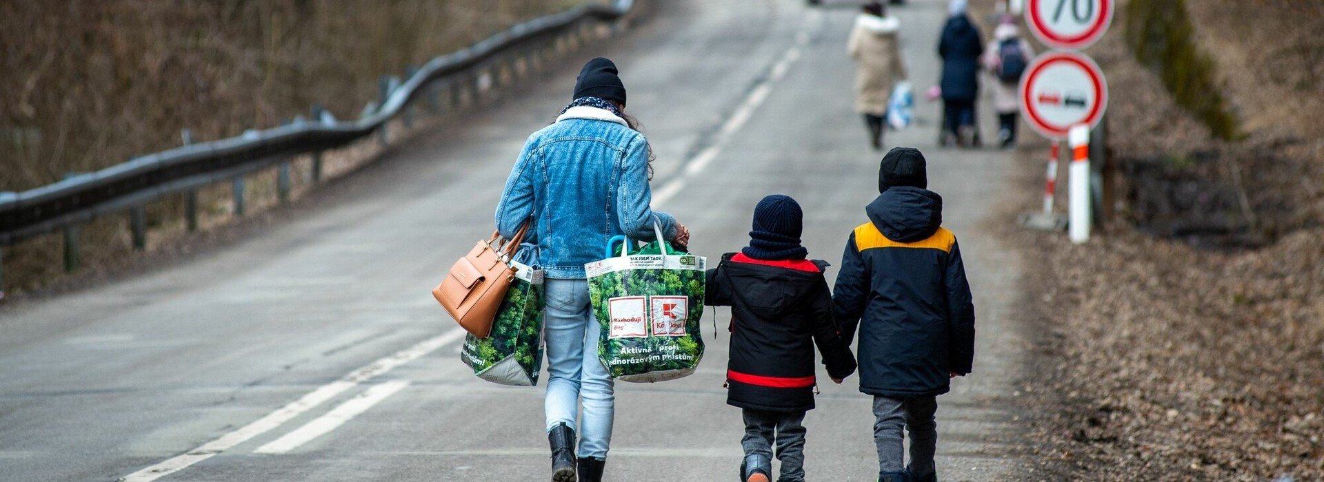 A mother and her two children carrying bags and walking on a road.