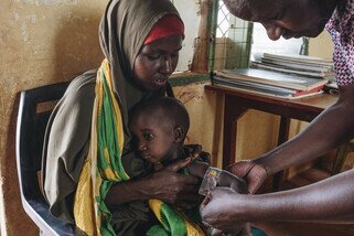 Mother holding her young child as a man measures the child's arm with a mid-upper arm circumference measuring tape.