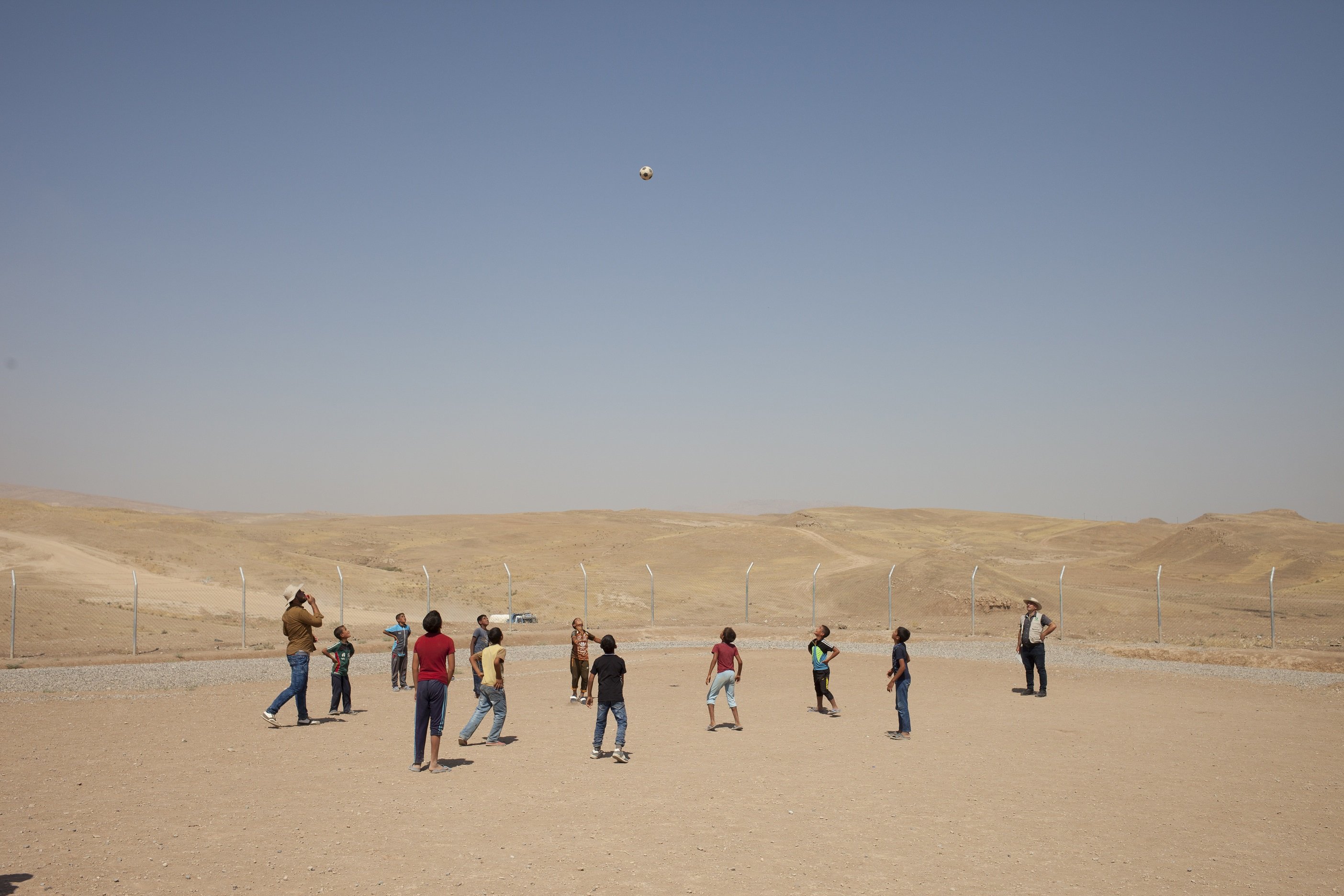 Children and adults playing soccer in a large open desert-like area.
