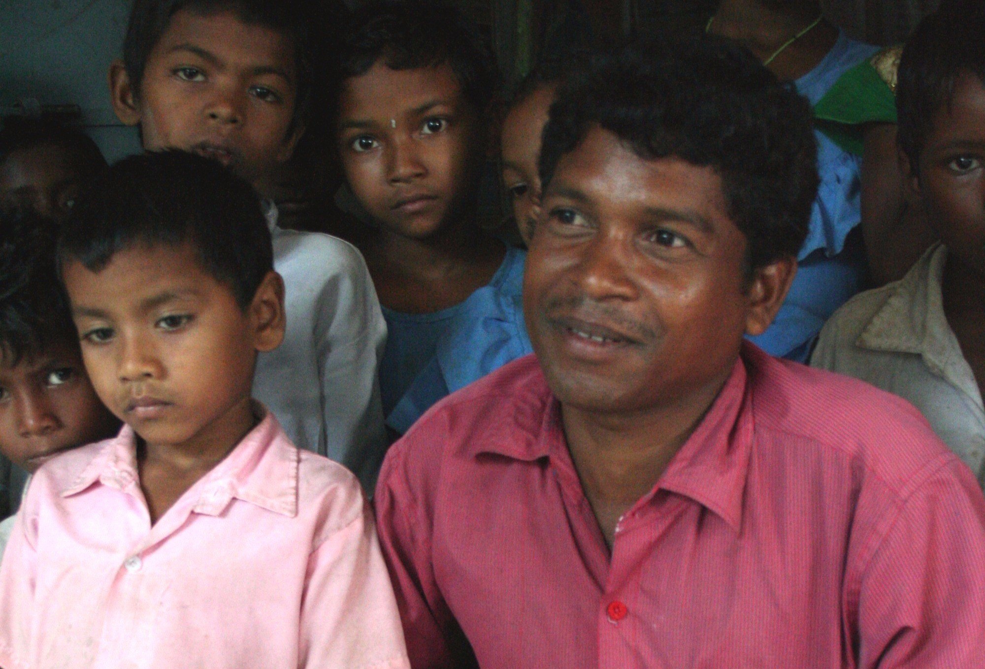 A man in a red shirt smiling. Several children surround him.