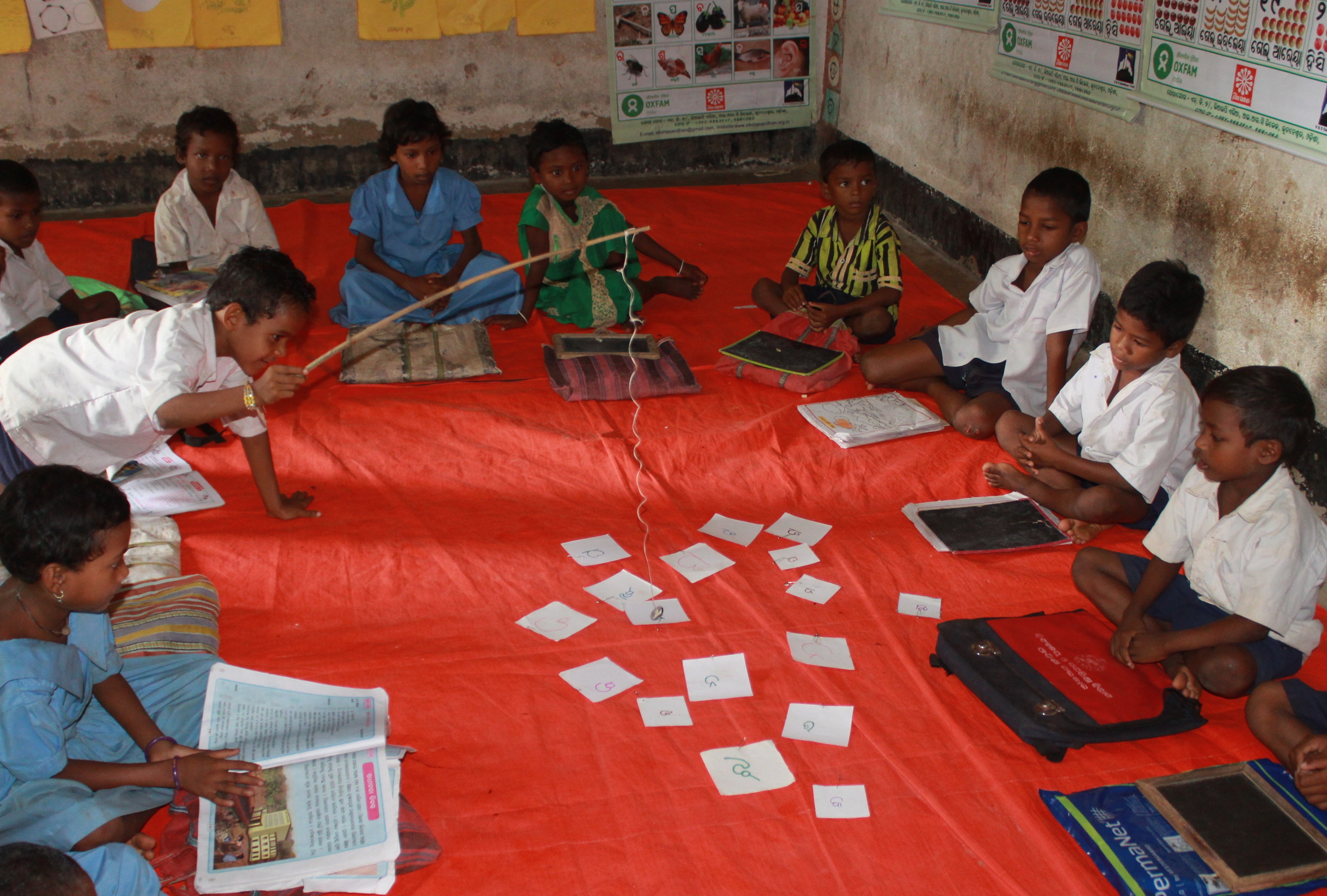 Children sitting in a circle on the floor in a classroom. One child is holding a stick and trying to pick up a letter in the Odia 'alphabet' as part of a game.
