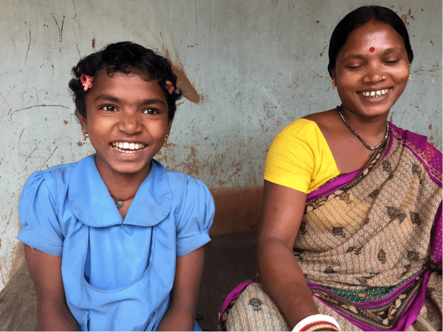 A young girl from Odisha, India, in her school uniform sitting next to her mother, who's wearing a sari.